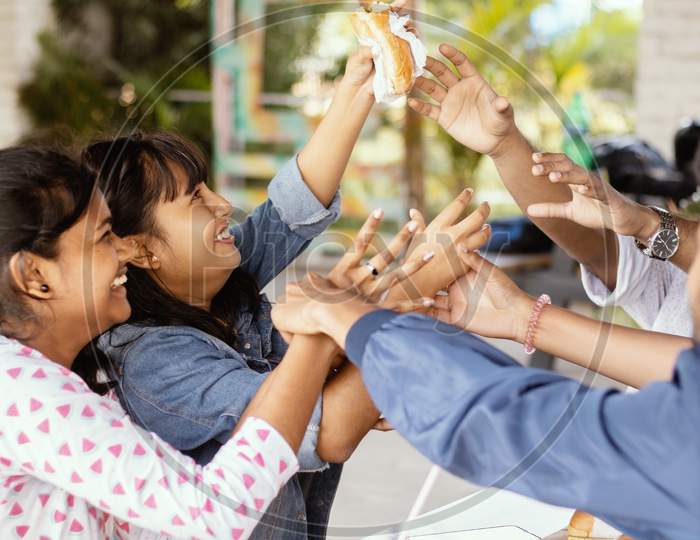 Group Of Friends Having Fun While Eating At Restaurant - All Friends Playfully Trying To Steal, Grab Or Taking Food From One Girls Hand - Teenager Students Having Fun While Having Food At College Cafeteria.