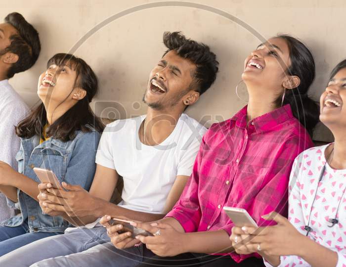 Group Of Happy Young Boys and Girls By Looking At Mobile Phone Laughing Loudly At University Campus - Millennials Enjoying Online Video Content Or Social Media By Watching Smart Phone.