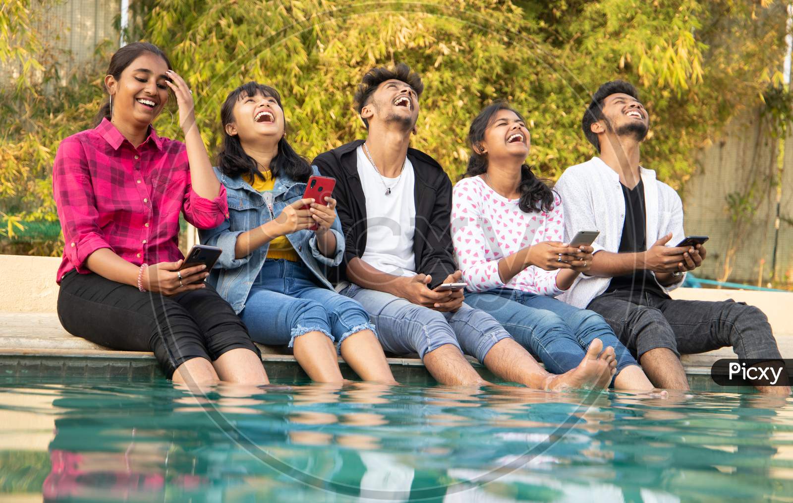 Group Of Happy Young Boys and Girls By Looking At Mobile Phone Laughing Loudly - Millennials Enjoying Online Video Content Or Social Media By Watching Smart Phone.