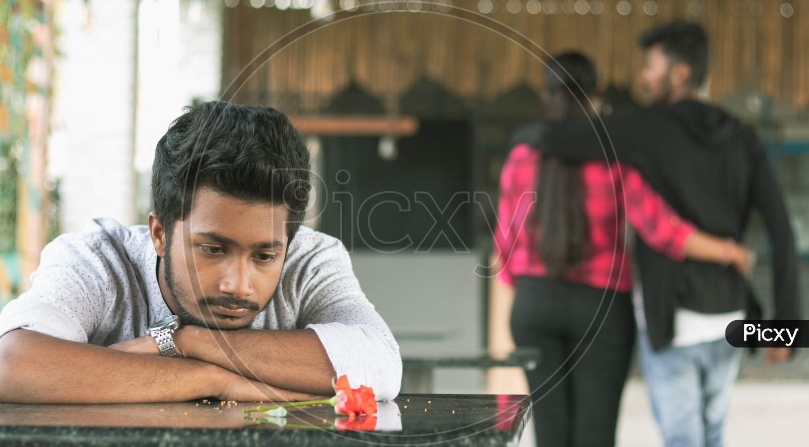 Selective Focus On Red Rose Petals, Lonely Young Teenager Sitting Sadly On Table By Laying his Head Down - Concept Of Love Breakup Or Broken Heart.
