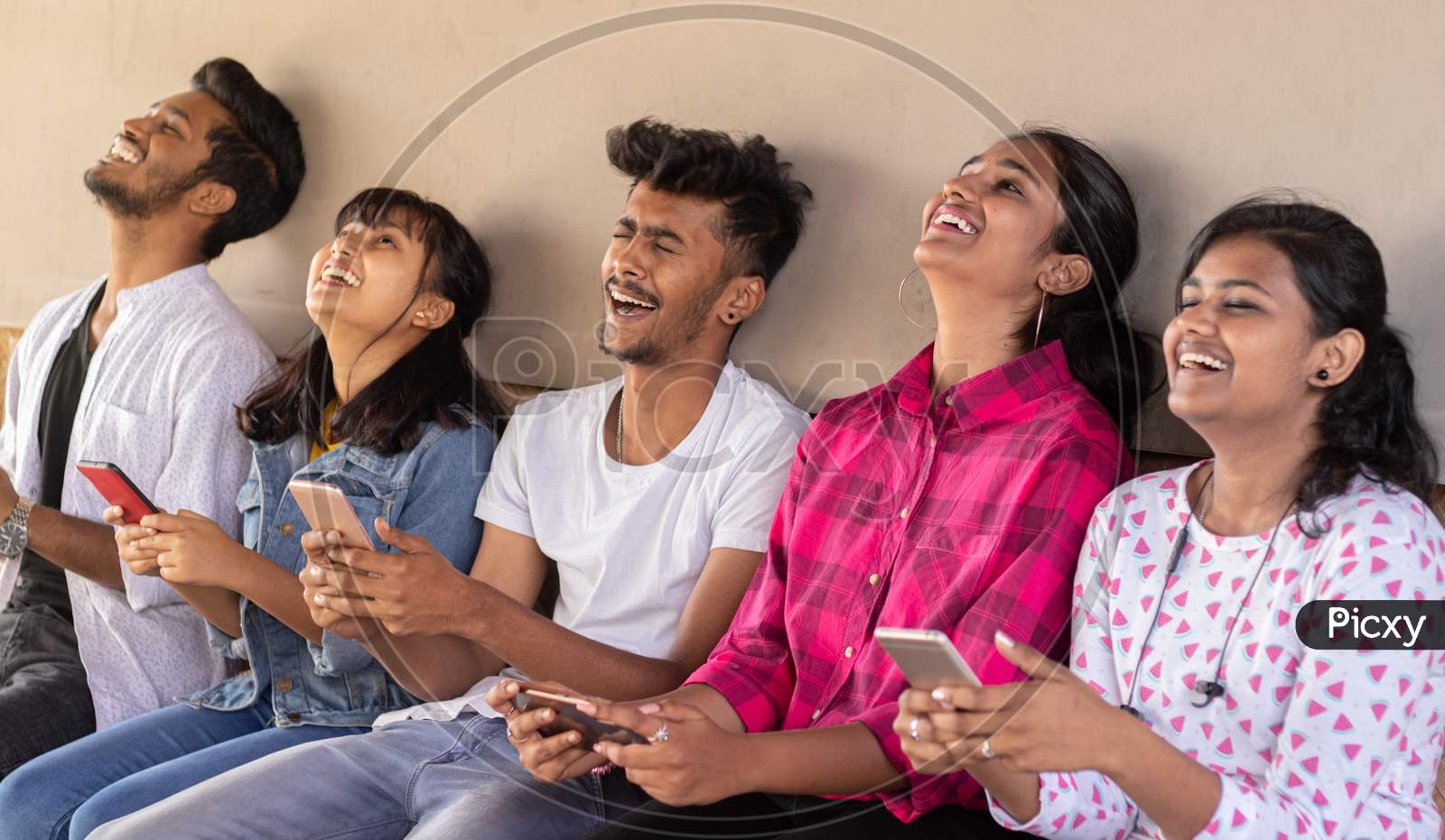 Group Of Happy Young Boys and Girls By Looking At Mobile Phone Laughing Loudly At University Campus - Millennials Enjoying Online Video Content Or Social Media By Watching Smart Phone.