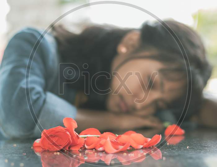 Selective Focus On Red Rose Petals, Lonely Young Teenager Sitting Sadly On Table By Laying Her Head Down - Concept Of Love Breakup Or Broken Heart.