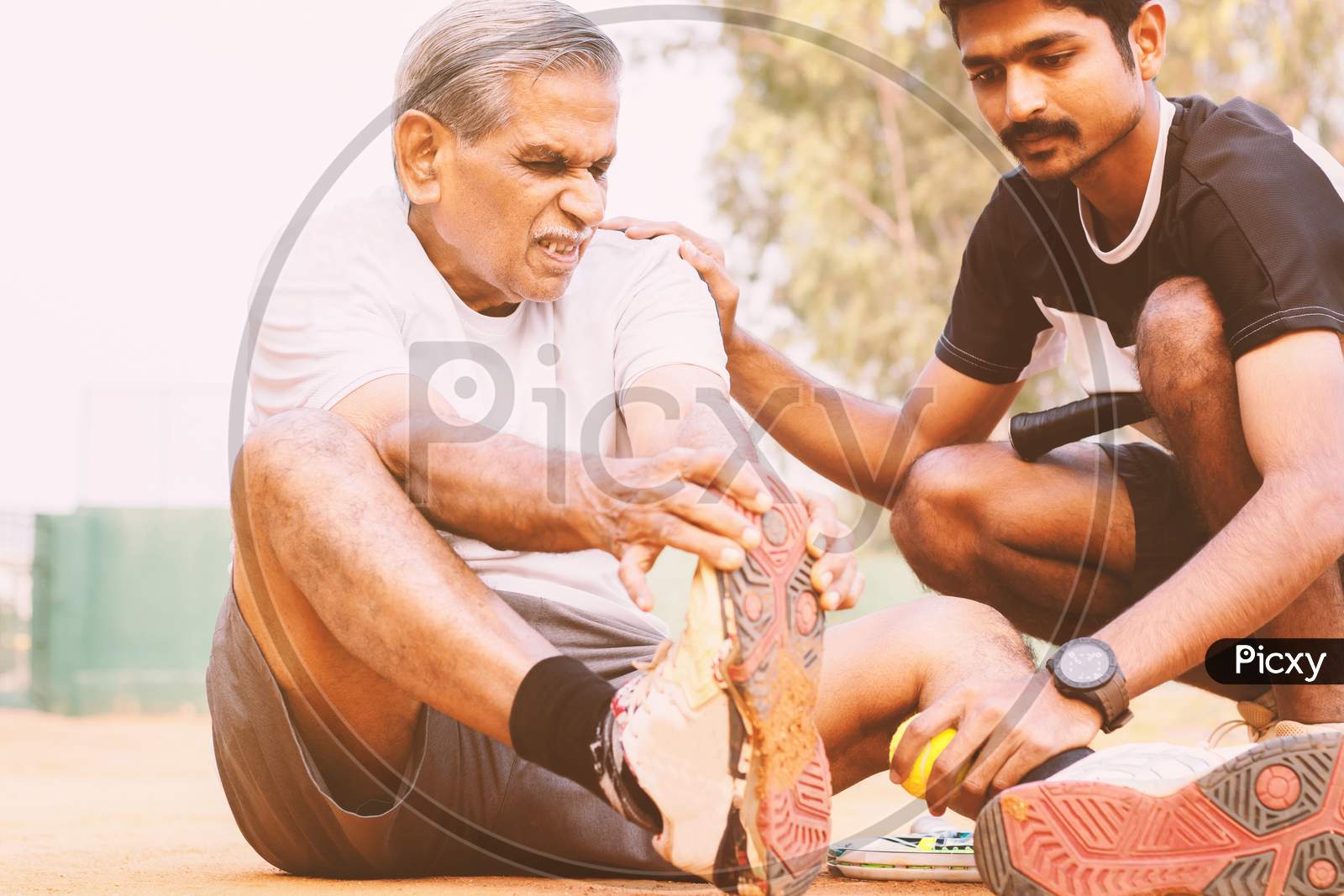 Elderly Holding His Toe Suffering From Pain And Young Man Or Teammate Helping Him At Tennis Court - Concept Of Old People Fitness And Injury - Young People Helping Senior Citizens At Sports.