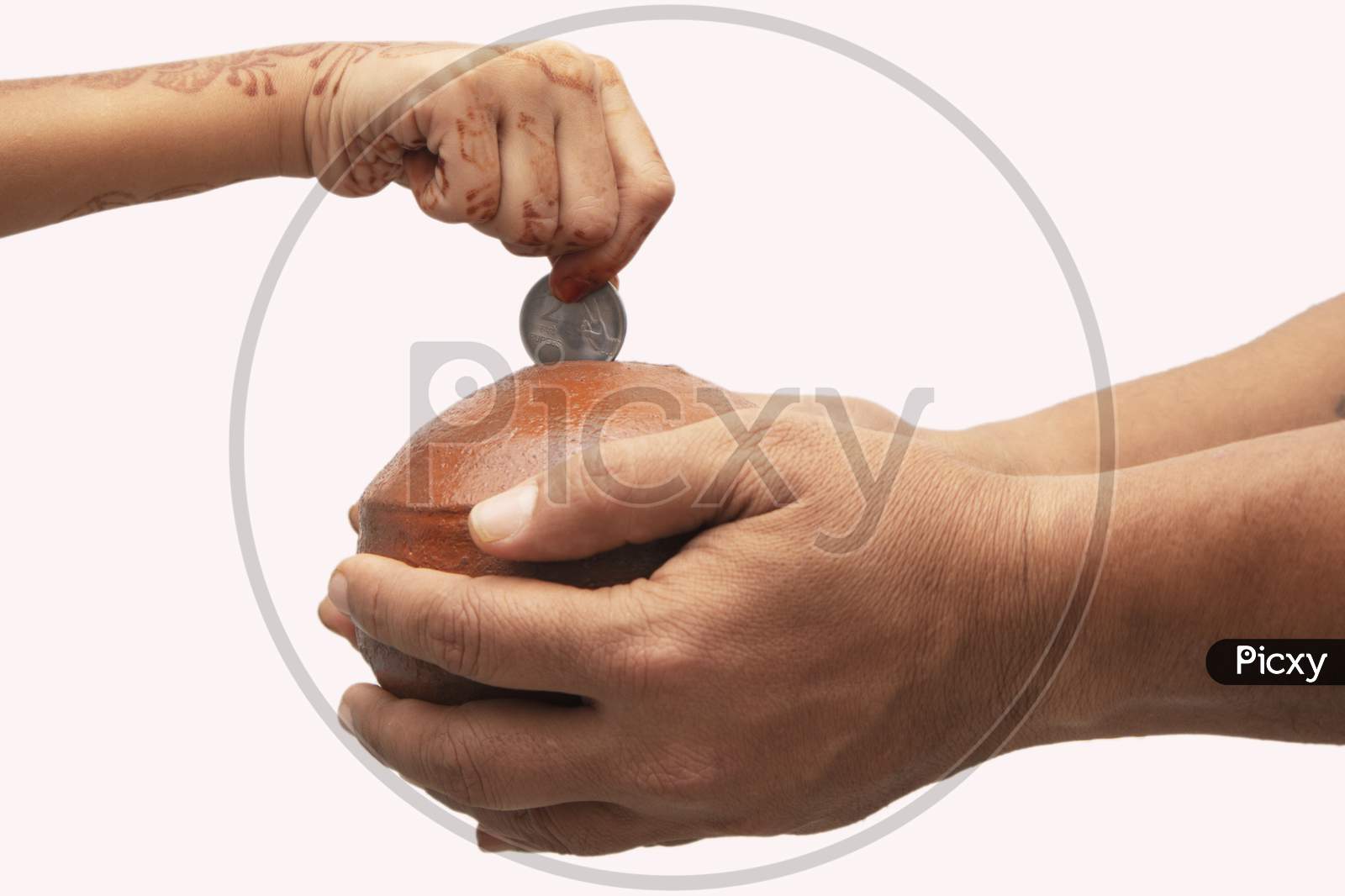 Hands Holding Indian Penny Bank Made With Mud Clay And Child Hand Adding Coin Into The Earthen Pot Or Terracotta Piggy Bank On Isolated Background - Concept Showing Of Saving, Investment Or Donating.