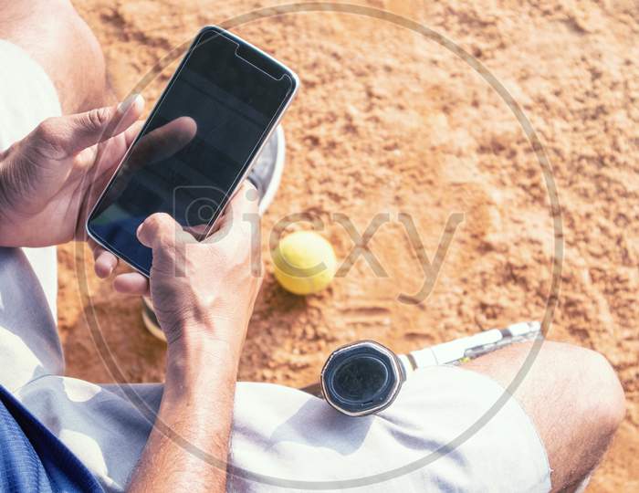 High Angle Shot Of Young Tennis Player Taking Rest After Training And Busy In Using Mobile Phone - Fitness, Sport And Recreation Concept