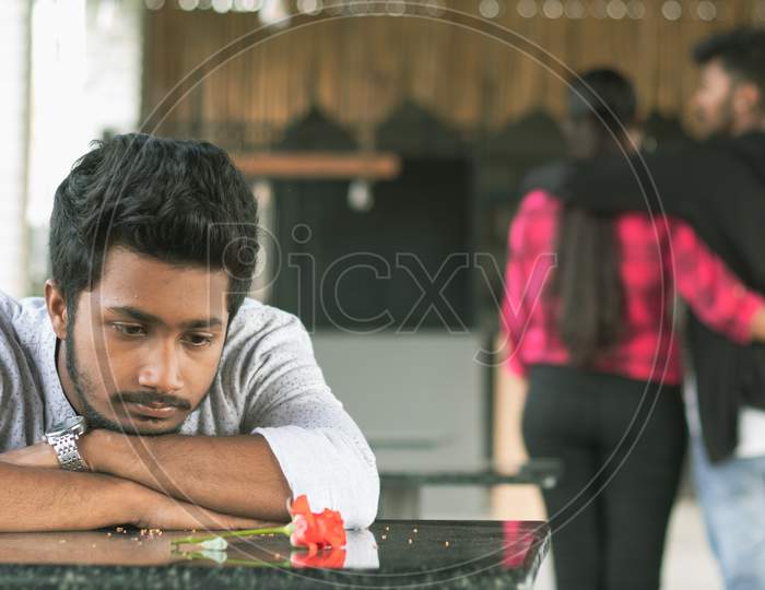 Selective Focus On Red Rose Petals, Lonely Young Teenager Sitting Sadly On Table By Laying his Head Down - Concept Of Love Breakup Or Broken Heart.