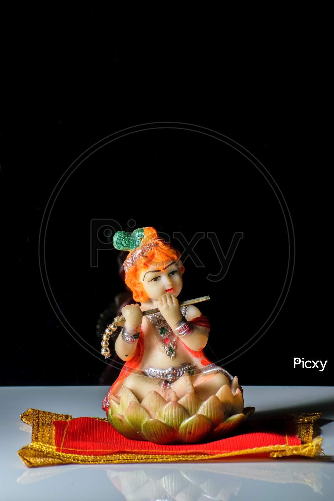 An Isolated Sculpture Of Indian God Lord Krishna Playing Flute Sitting On A Mat. On A Reflective White Table With A Dark Background.Portrait View