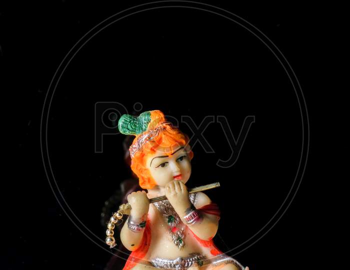 An Isolated Sculpture Of Indian God Lord Krishna Playing Flute Sitting On A Mat. On A Reflective White Table With A Dark Background.Portrait View