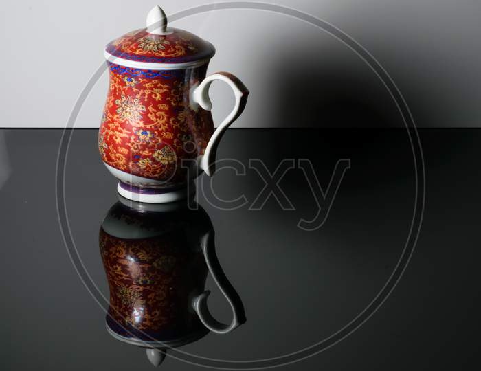A Red Teapot With Design On It Placed On A Reflective Table In A White Background