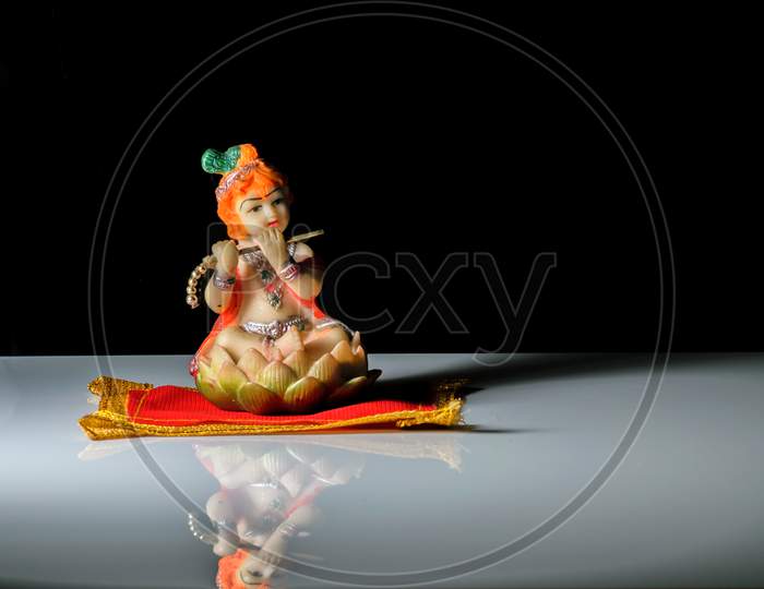 An Isolated Sculpture Of Indian God Lord Krishna Playing Flute Sitting On A Mat. On A Reflective White Table With A Dark Background