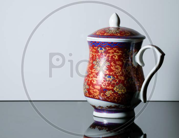 A Red Teapot With Design On It Placed On A Reflective Table In A White Background