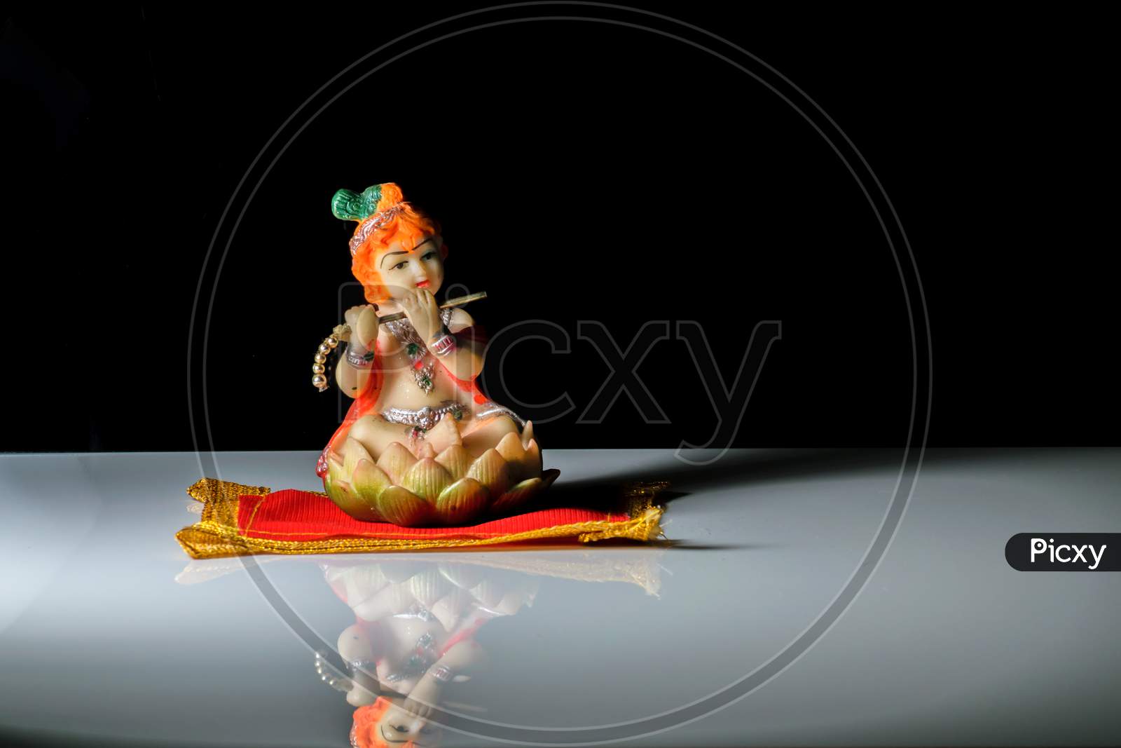An Isolated Sculpture Of Indian God Lord Krishna Playing Flute Sitting On A Mat. On A Reflective White Table With A Dark Background