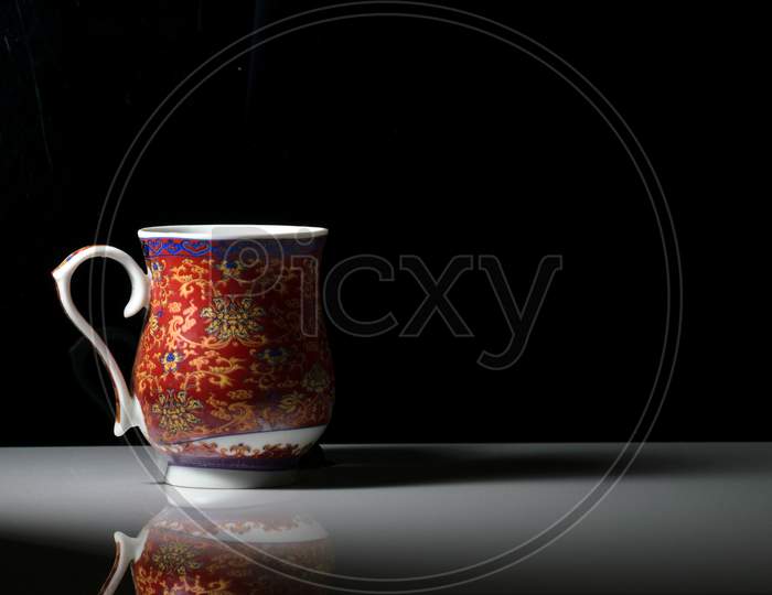 A Red Tea Cup With Designs On It Placed On A White Reflective Table And Smokes Of Hot Tea Coming Out Of It. Landscape View