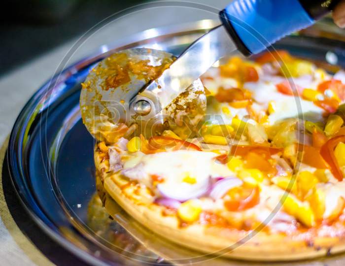 Cutting cheese pizza with Pizza cutter, with tomato, onion, corn toppings, with selective focus in lockdown.