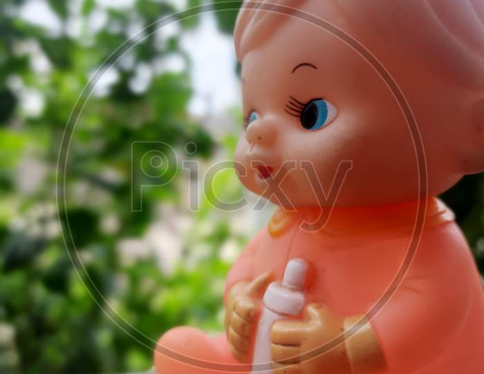 a baby toy with blue eyes small nose and mouth having a bottle of milk holded in hands in blur greenish background