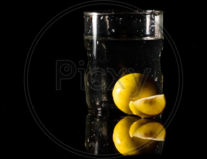 A Glass Of Water Placed On A Reflective Surface In A Dark Background With Cut And Intact Lemon Placed In Front Of It