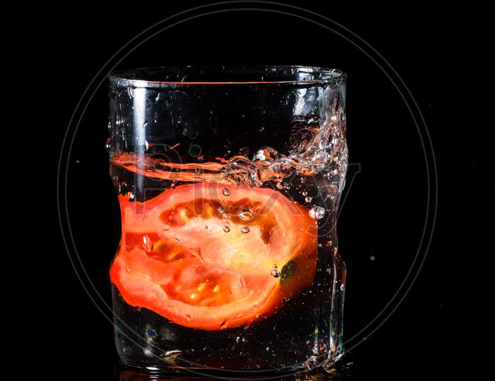 A Glass Of Water In A Dark Background And Water Being Splashed With A Piece Of Cut Tomato Dropped Inside The Glass