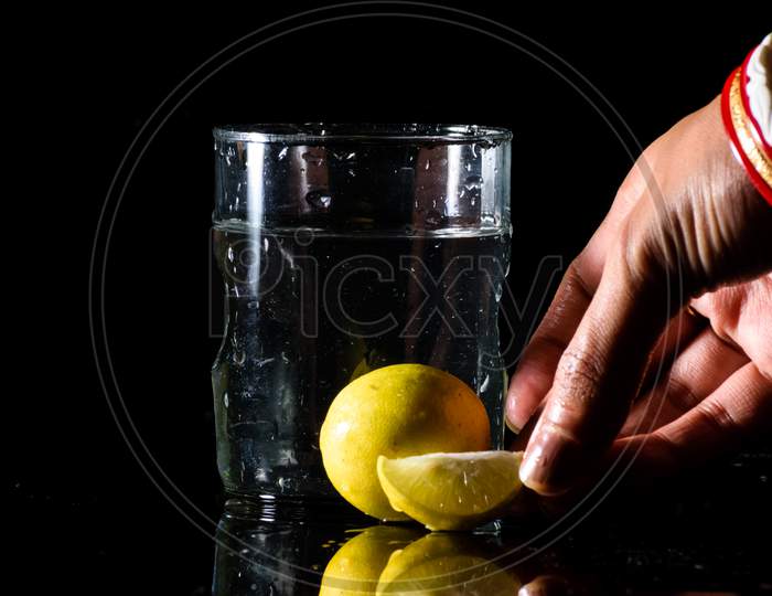 A Glass Of Water Placed On A Reflective Surface In A Dark Background With Cut And Intact Lemon Placed In Front Of It And Being Held By A Hand