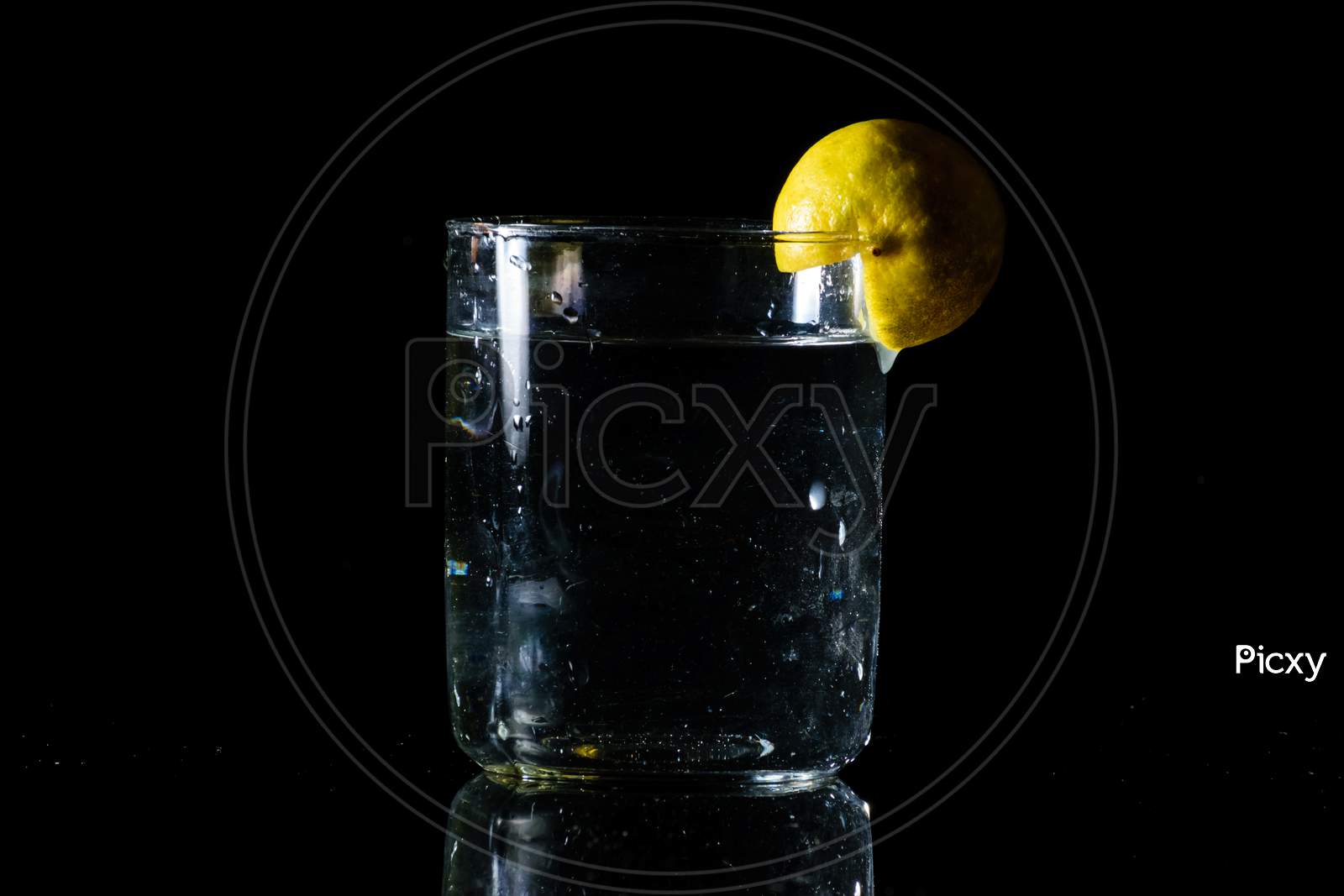 A Glass Of Water Placed On A Reflective Surface In A Dark Background And A Cut Lemon On The Edge Of The Glass