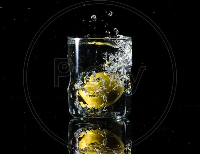 A Glass Of Water Placed On A Reflective Surface With Black Background And Water Is Splashing After Dropping A Lemon In The Glass. Summer Concept