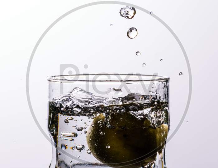 A Glass Of Water Placed On A Reflective Surface With White Background And Water Is Splashing After Dropping A Lemon In The Glass