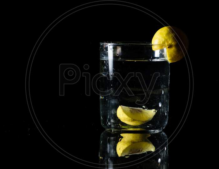 A Glass Of Water Placed On A Reflective Surface In A Dark Background And A Cut Lemon Inside And On The Edge Of The Glass