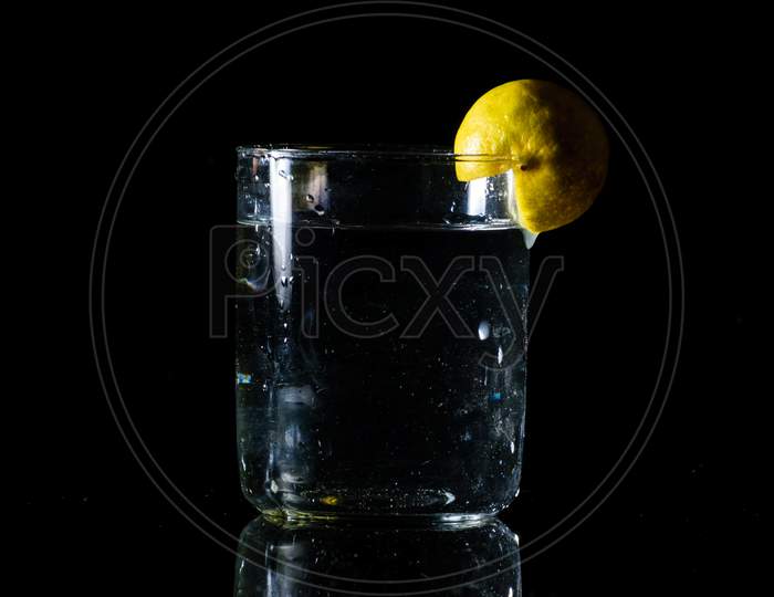 A Glass Of Water Placed On A Reflective Surface In A Dark Background And A Cut Lemon On The Edge Of The Glass