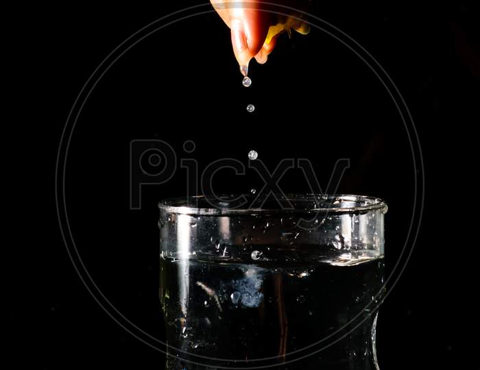 A Glass Of Water Placed On A Reflective Surface In A Dark Background And A Piece Of Cut Lemon Is Being Squeezed From Above And The Juice Is Falling