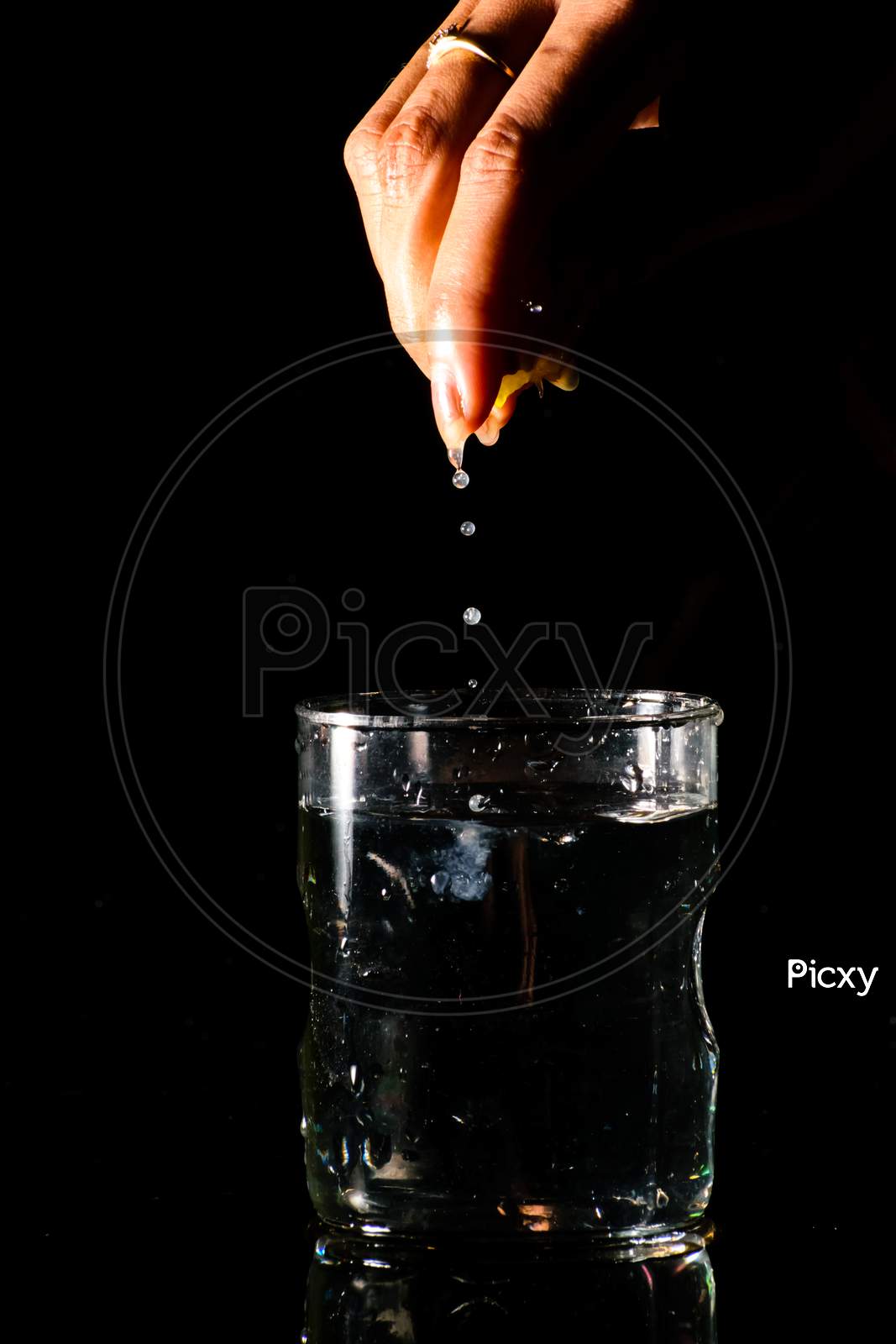 A Glass Of Water Placed On A Reflective Surface In A Dark Background And A Piece Of Cut Lemon Is Being Squeezed From Above And The Juice Is Falling