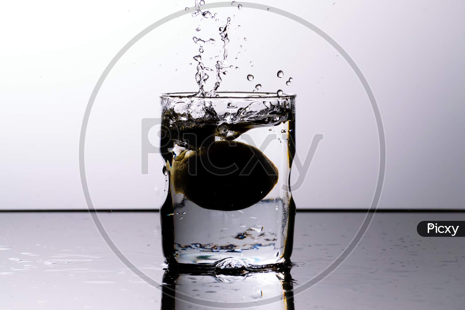A Glass Of Water Placed On A Reflective Surface With White Background And Water Is Splashing After Dropping A Lemon In The Glass