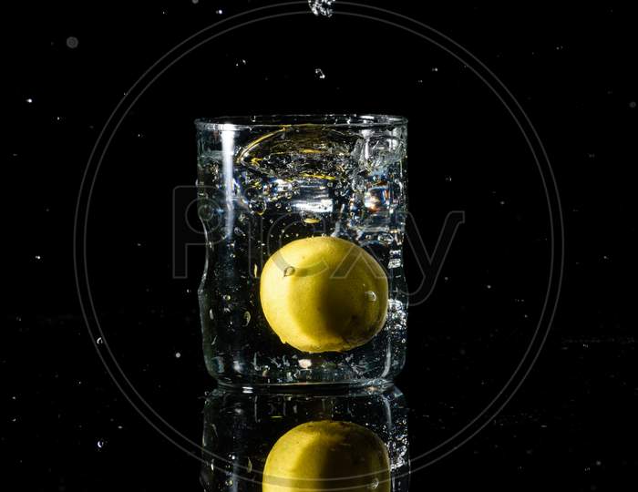 A Glass Of Water Placed On A Reflective Surface With Black Background And Water Is Splashing After Dropping A Lemon In The Glass. Summer Concept