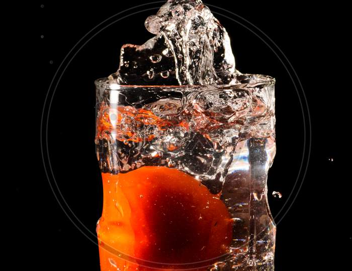 A Glass Of Water In A Dark Background And Water Being Splashed With A Piece Of Cut Tomato Dropped Inside The Glass