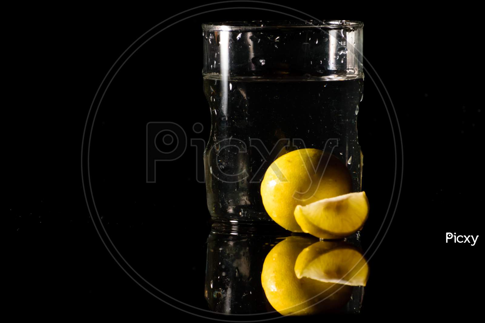 A Glass Of Water Placed On A Reflective Surface In A Dark Background With Cut And Intact Lemon Placed In Front Of It