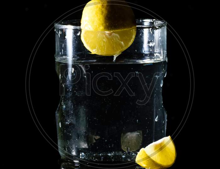 A Glass Of Water Placed On A Reflective Surface In A Dark Background With A Cut Lemon Placed In Front Of It And An Intact Lemon On The Edge Of The Glass
