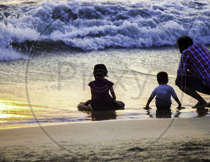 A Family Of A Father, Son And Daughter Sit On A Beach Against Waves In Sunset
