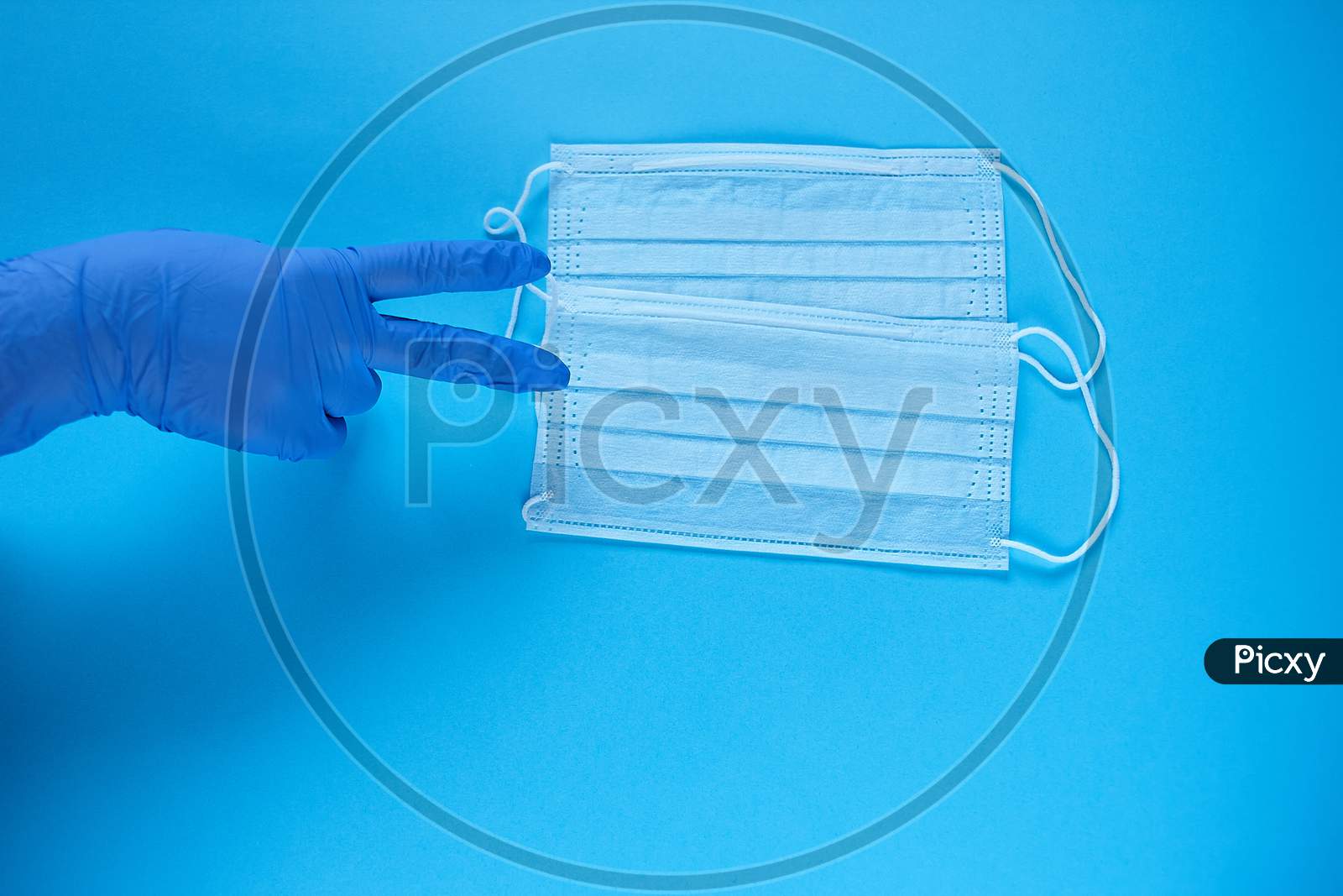 A Hand In Protective Medical Glove Points A Fingers At Two Protective Medical Masks. Protect From Coronavirus For Two People Concept On Blue Background