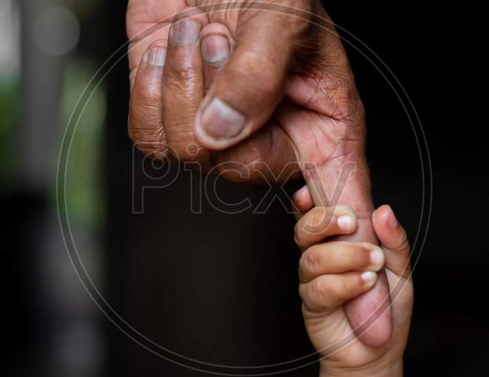 A Baby'S Hands Holding Tightly A Senior Man'S Old Age Finger. Family, Generation, Support And People Concept. Dark Background.