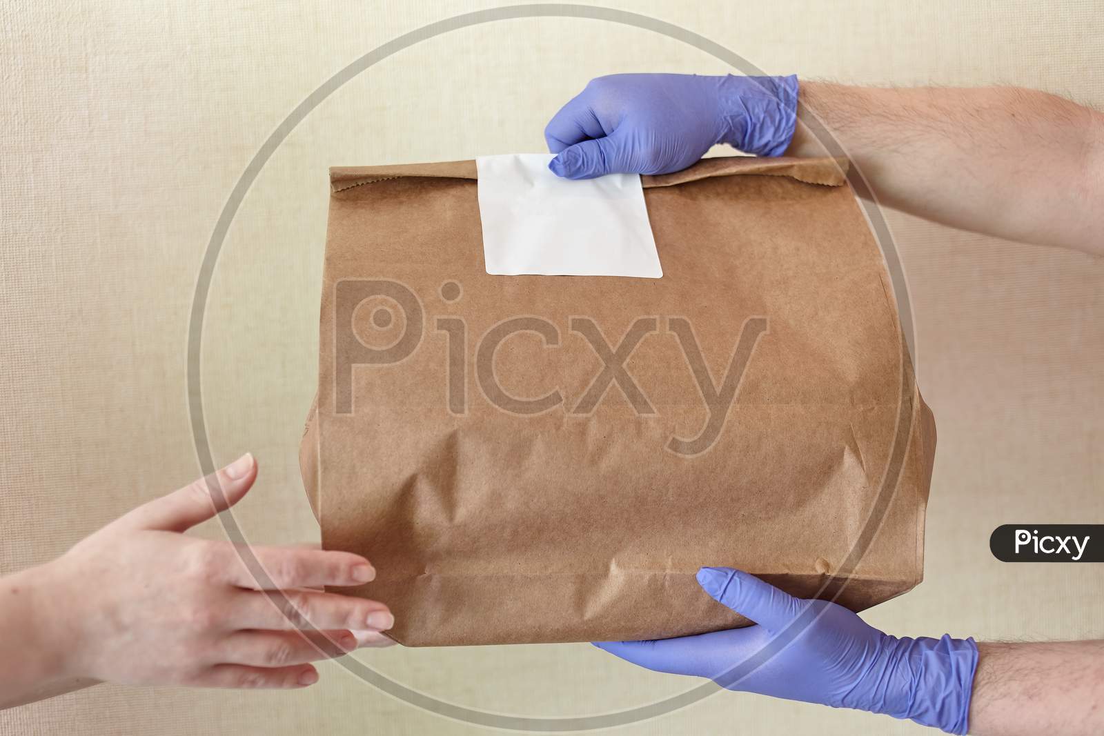 Male Hands In Blue Latex Glove Gives Paper Bag To Female Hands. Closeup Of Male Hands In Gloves Hold Brown Craft Paper Bag With Blank White Sticker. Delivery Service Concept. Copy Space Area.