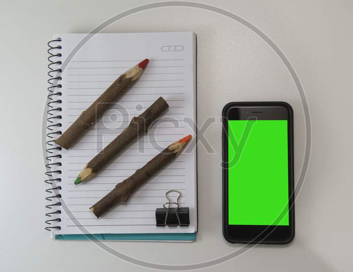 Top View Of Office Desk With Blank Note Book, Rustic Colored Pencils And Smartphone.