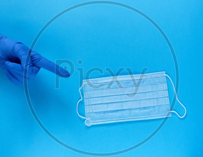A Hand In A Protective Medical Glove Points A Finger At A Protective Medical Mask. Typical Surgical Mask For Covering The Mouth And Nose. Protect From Coronavirus And Bacteria Concept On Blue Background