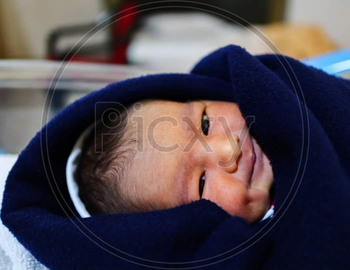 A Newborn Asian Baby Wrapped In Navy Blue Towel Looking At The Camera And Smiling With Dimple In Cheeks