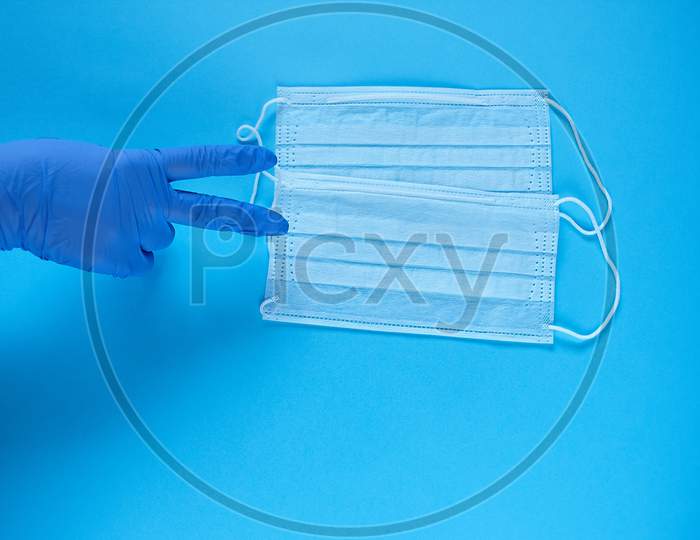 A Hand In Protective Medical Glove Points A Fingers At Two Protective Medical Masks. Protect From Coronavirus For Two People Concept On Blue Background
