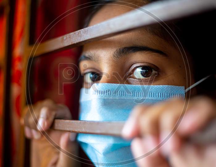 A Bored Asian Young Girl Wearing A Protection Surgical Face Mask At Looking Through Window Being In-Home Quarantine During Coronavirus Outbreak. Close-Up Views.