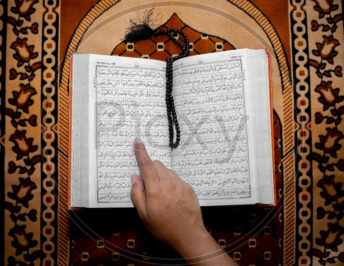 Muslim Woman Reading The Holy Quran Using The Finger. The Holy Quran On The Mat Of Prayers . Top Angle Views. Indoors.