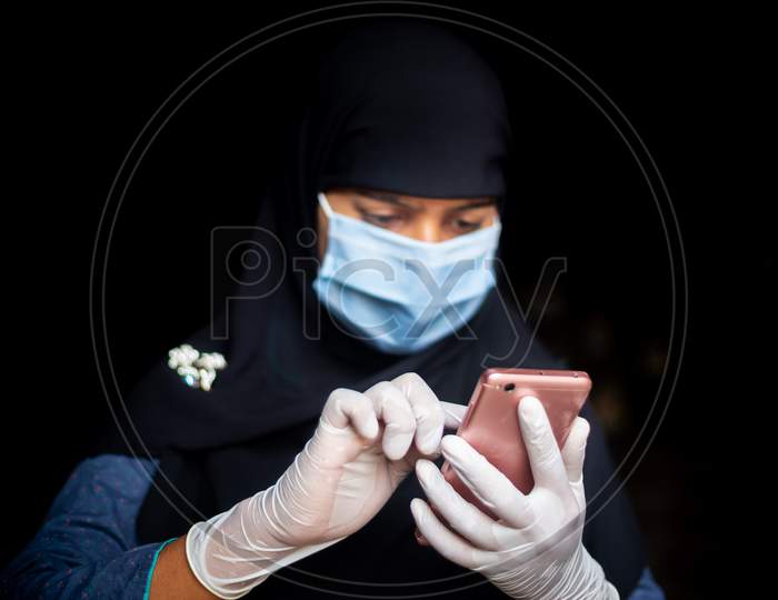 A Safety Mask And Gloves-Wearing A Muslim Girl Is Browsing His Smartphone For Coronavirus News. Black Hijab Woman Wearing A Blue Mask For Coronavirus Safety.
