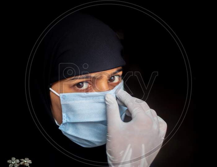 Muslim Girl Adjusting A Surgical Mask For Coronavirus Protection. Black Hijab Woman Wearing A Blue Mask For Safety. Dark Background Views.