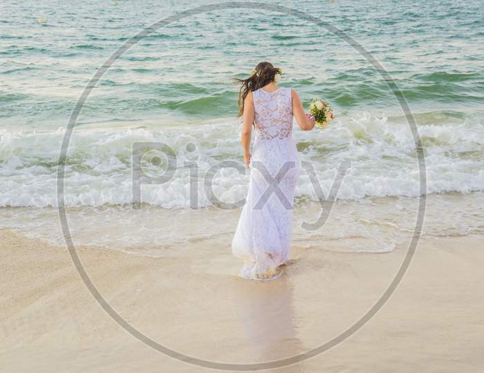Back View Of Bride Entering The Sea With Wedding Dress And Bouquet In Hand.