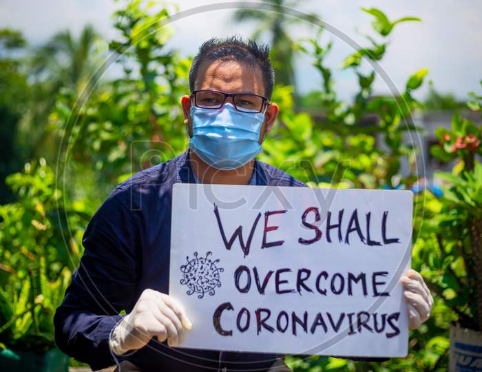 A Young Man Wearing A Medical Mask And Safety Gloves Stands With A Placard Message To "We Shall Overcome Coronavirus". A Man Holding A Placard Message In Outdoor Natural Light.