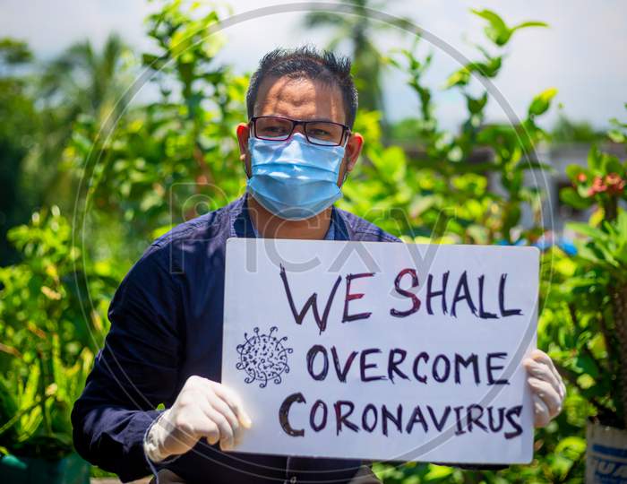 A Young Man Wearing A Medical Mask And Safety Gloves Stands With A Placard Message To "We Shall Overcome Coronavirus".A Man Holding A Placard Message In Outdoor Natural Light.