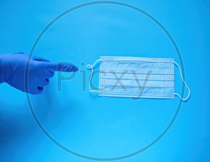 A Hand In A Protective Medical Glove Points A Finger At A Protective Medical Mask. Protect From Coronavirus Concept On Blue Background